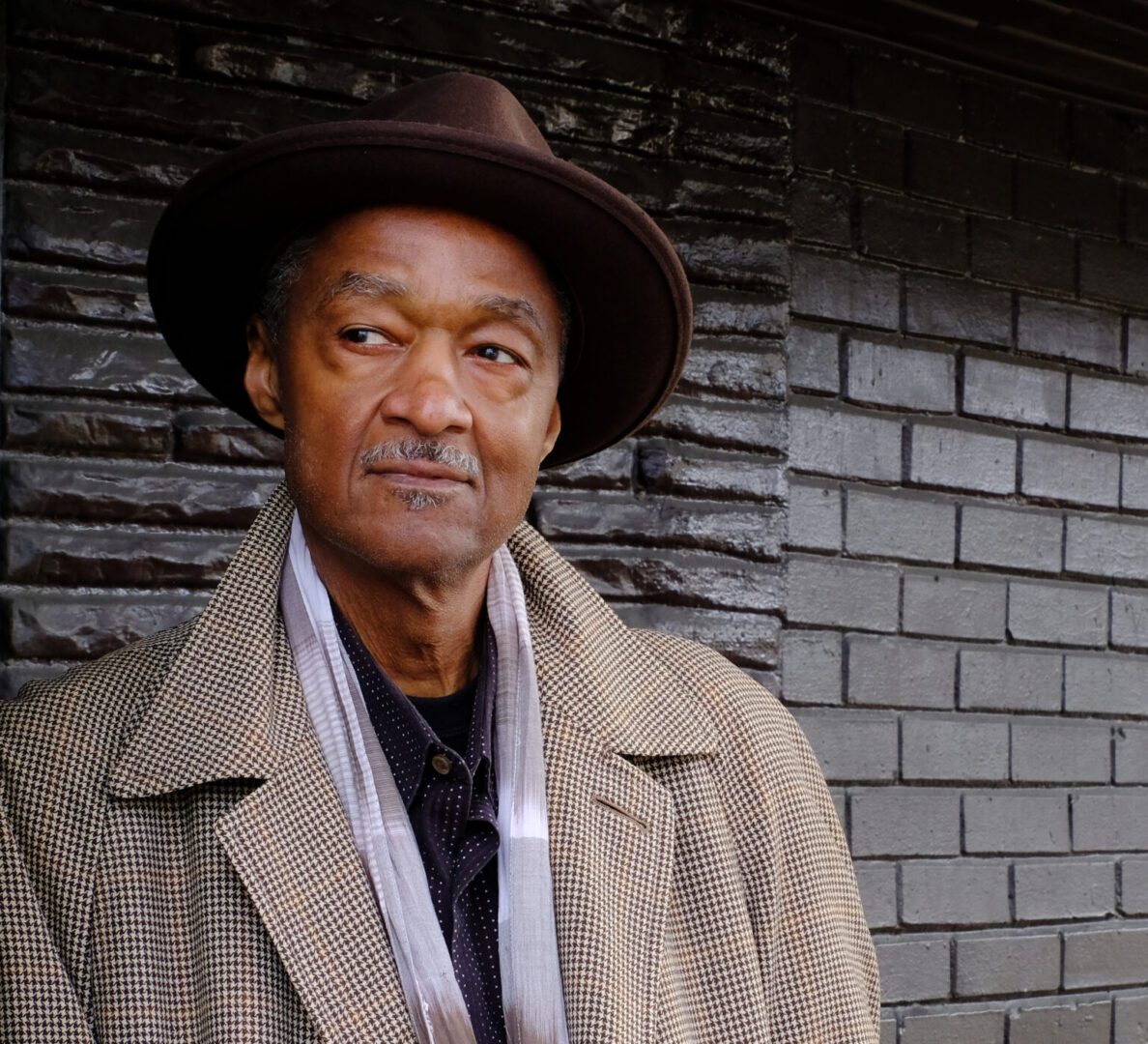 A man in a hat and coat standing next to a brick wall.