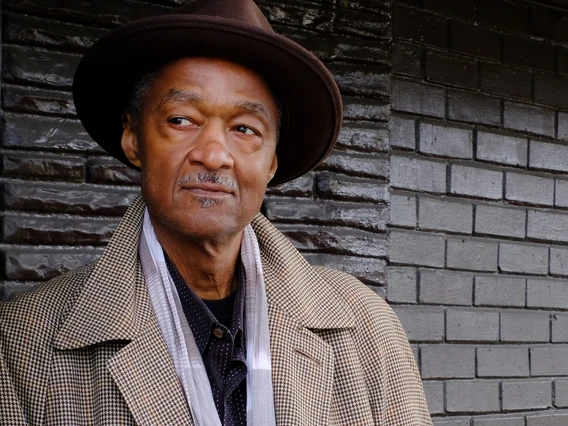 A man in a hat and jacket standing next to a brick wall.